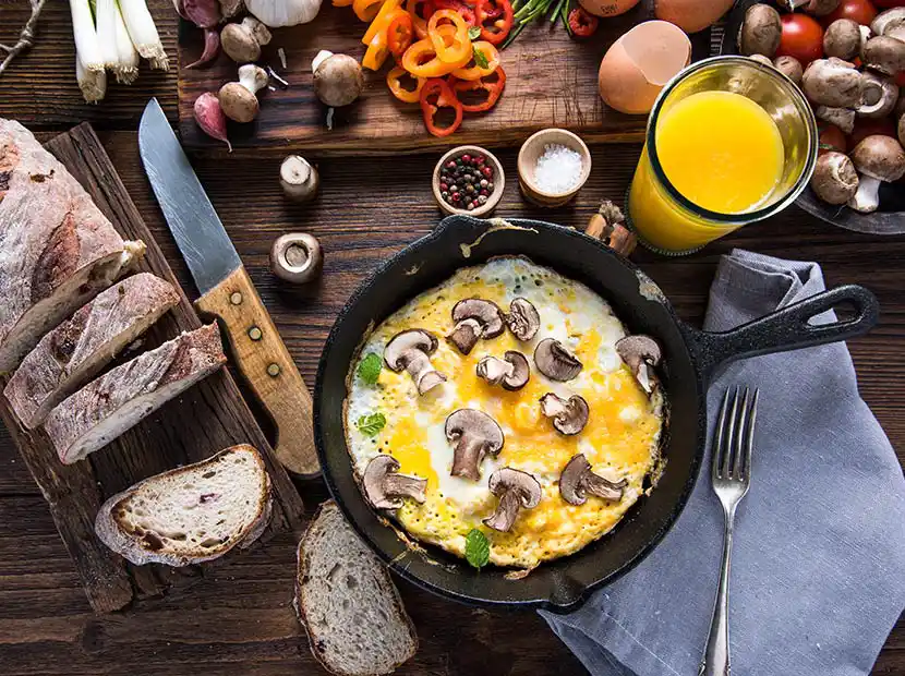 Pilzomlett in der Pfanne mit Orangensaft, Brot und Kaffee auf Holztisch.