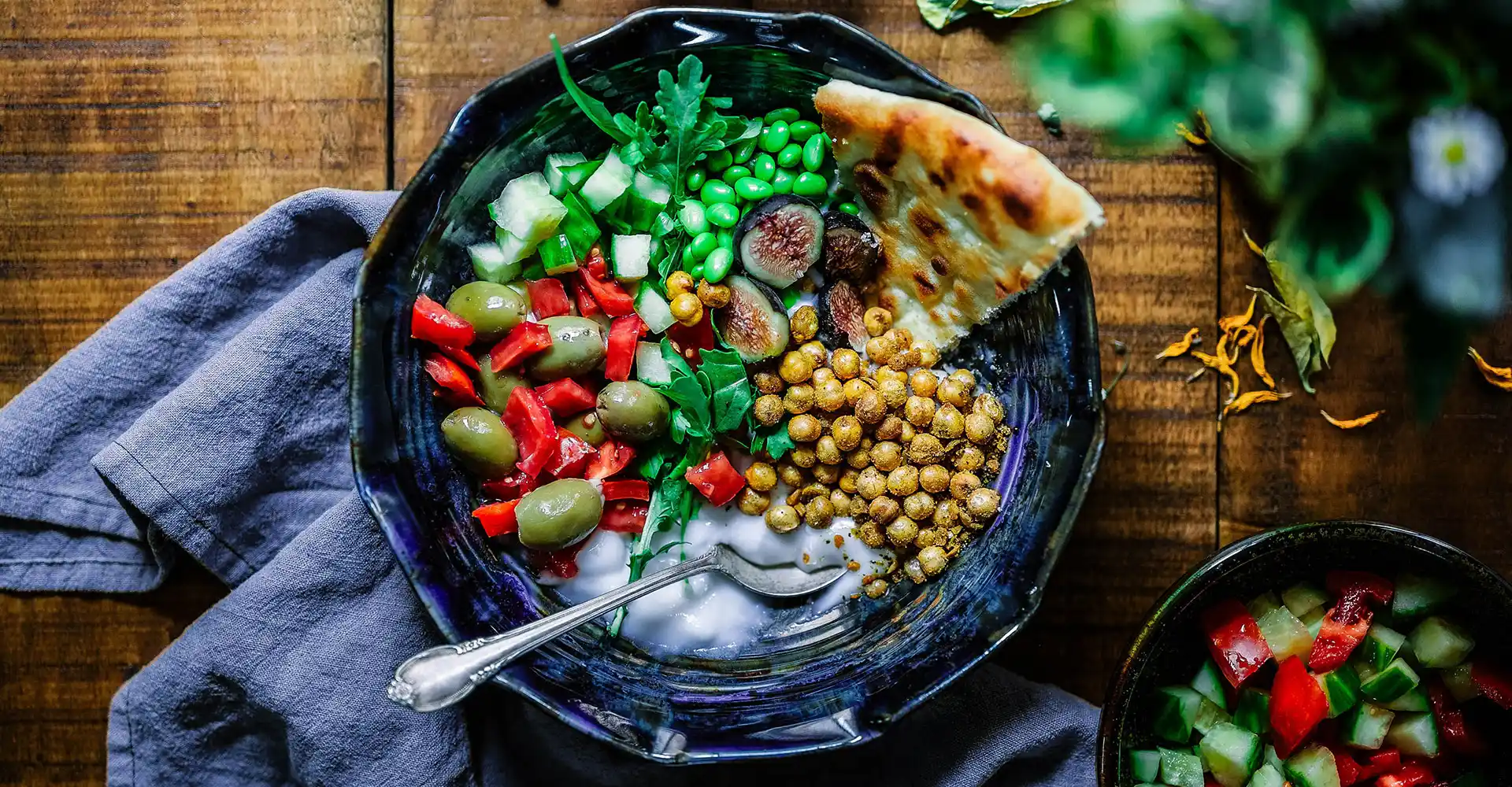 Salatschüssel auf Holztisch mit Gurken, Feigen, Oliven, Erbsen, Kichererbsen und Joghurt.