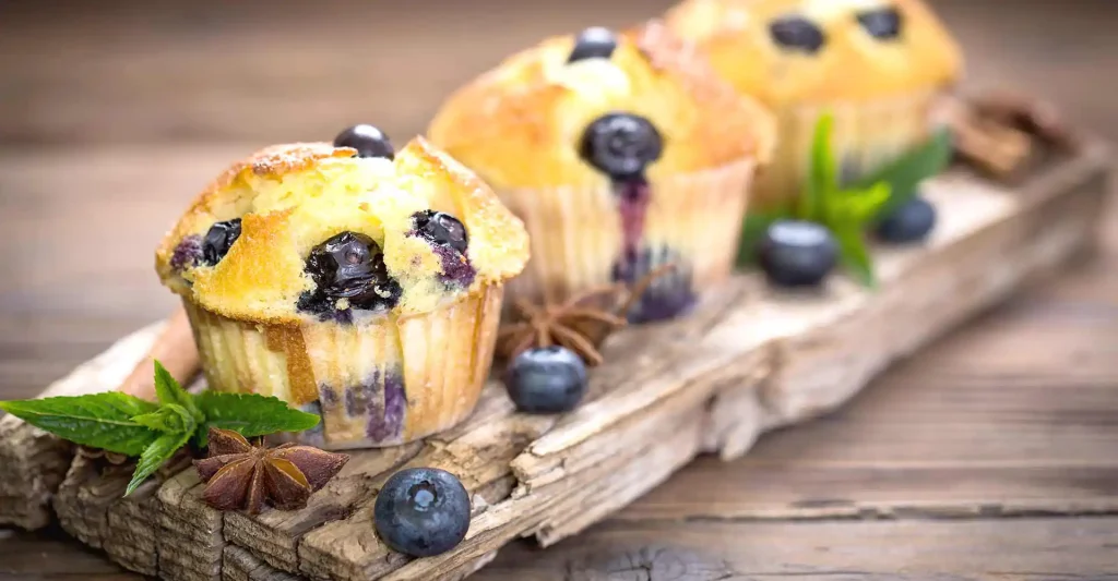Holzbrett mit 3 Blaubeer-Muffins und Minzgarnitur.
