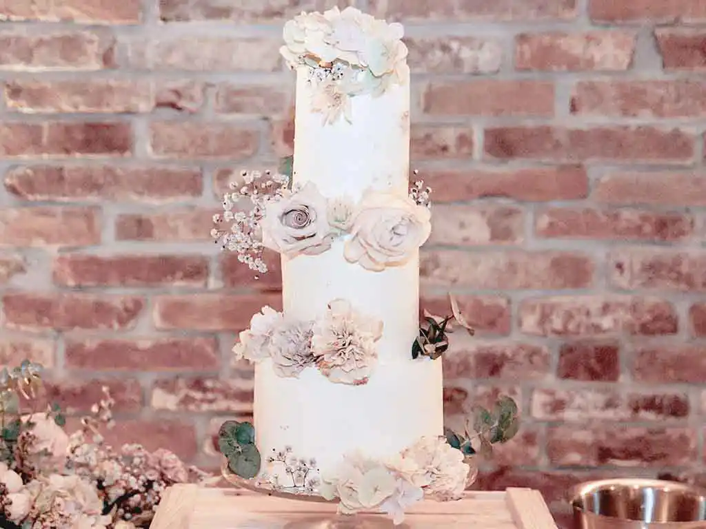 Tisch mit 3-stöckiger weißer Hochzeitstorte mit Blumendekor vor einer roten Ziegelwand.